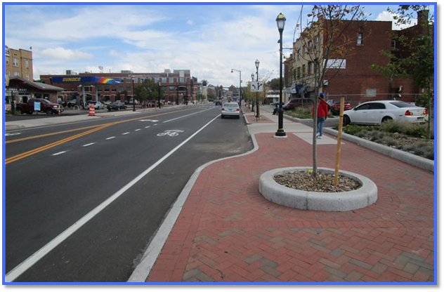 "A streetscape showing a newly renovated roadway with two travel lanes, a designated bike lane, and on‑street parking. A wide brick-paved sidewalk runs alongside the street with landscaped tree pits bordered by concrete edging. Buildings line both sides of the street, including storefronts and a gas station in the background. Streetlights and small trees are evenly spaced along the sidewalk, and a few vehicles are parked along the curb."